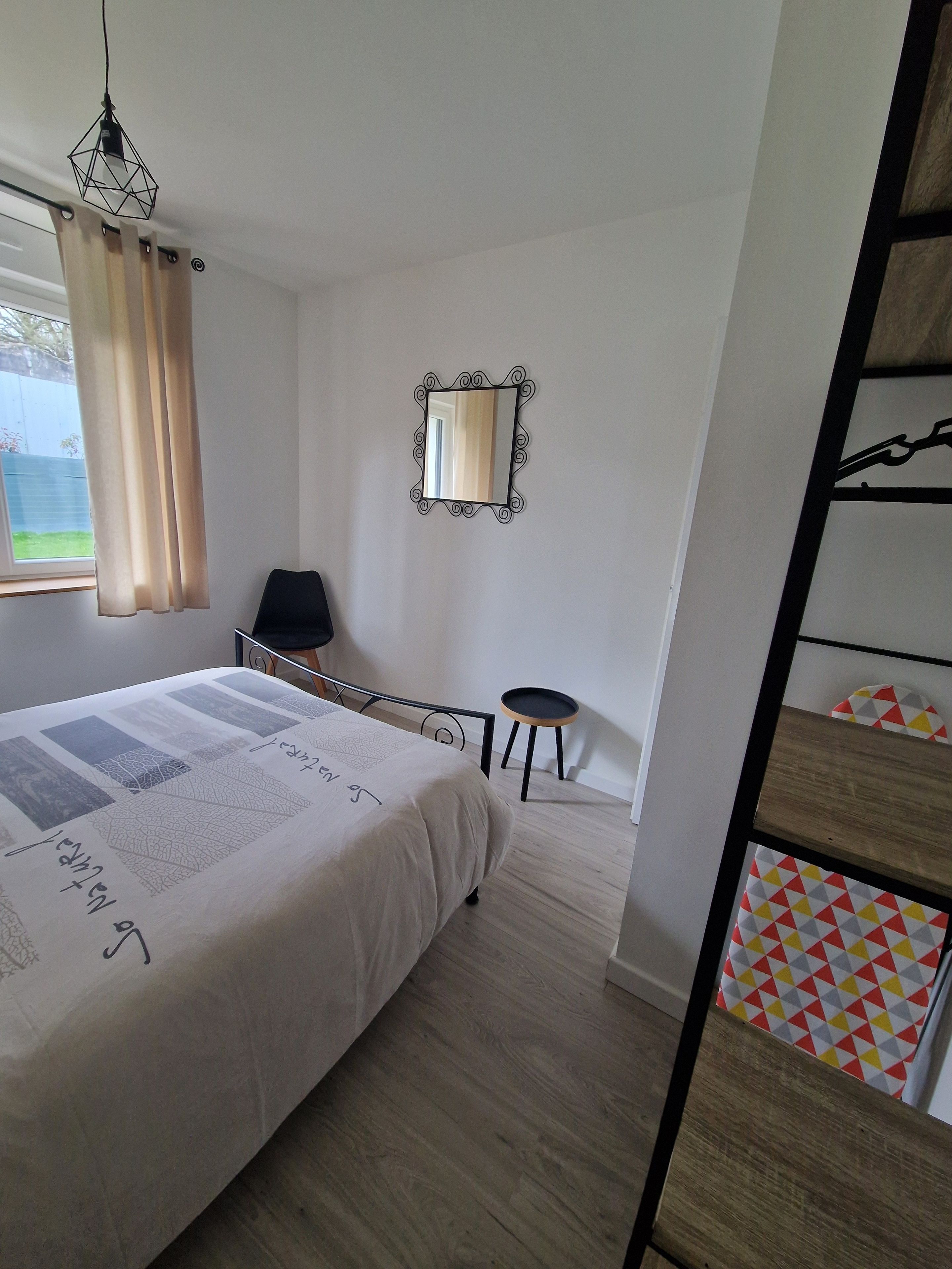 Une chambre à coucher avec un lit, une chaise, une petite table, un miroir et une fenêtre avec des rideaux.