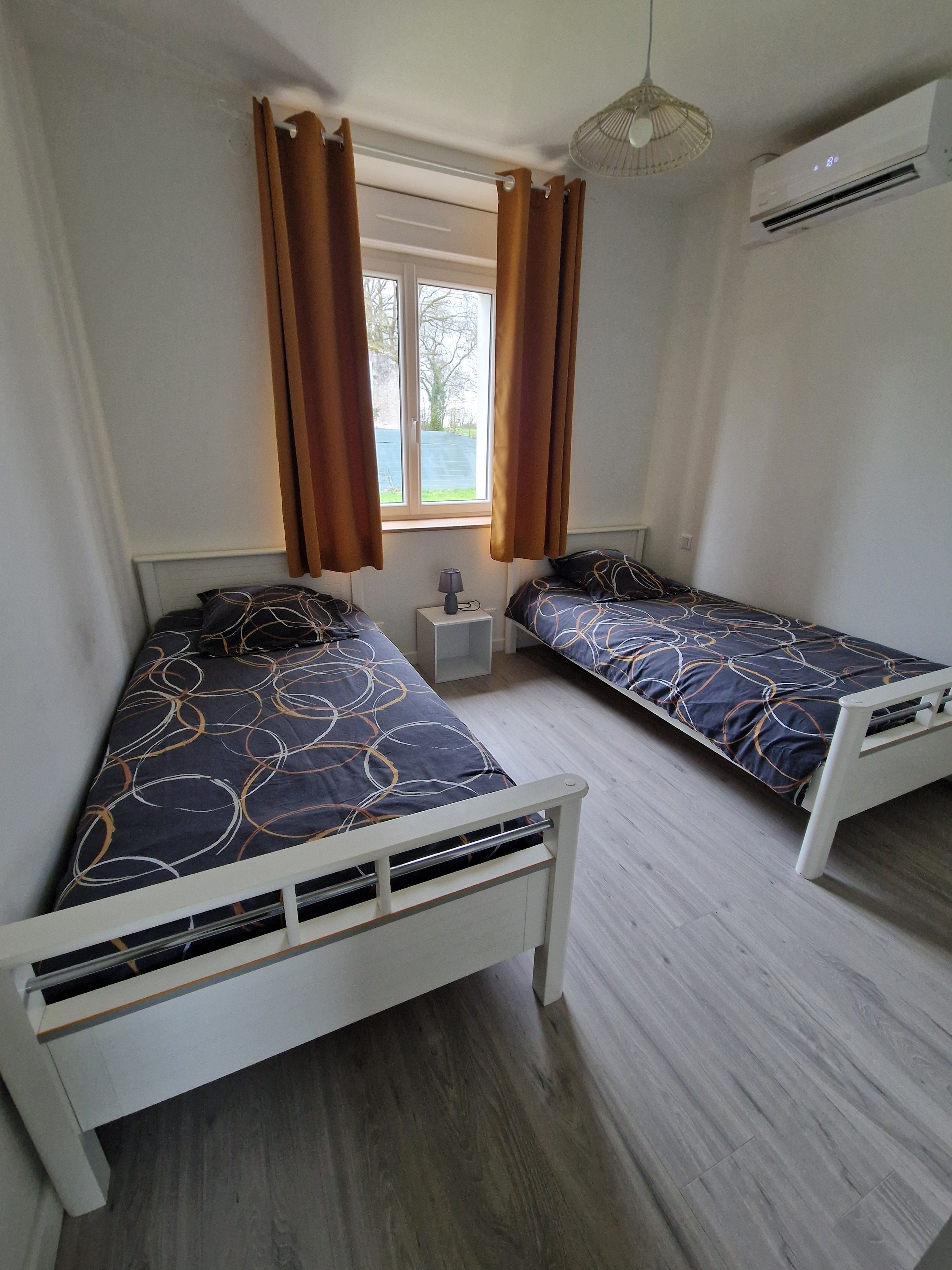 Habitació amb dos llits individuals, una finestra amb cortines, un aparell d'aire condicionat i una taula de nit amb una làmpada.