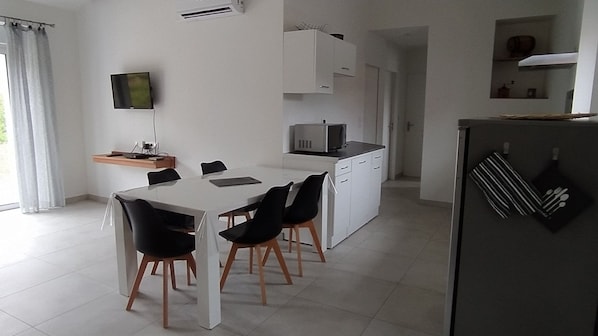 Een moderne keuken met witte kasten, een eettafel met stoelen, een aan de muur bevestigde televisie en een koelkast met keukengerei erop.