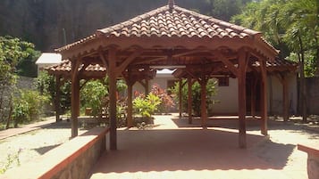 A wooden gazebo with a tiled roof, surrounded by plants and trees.
