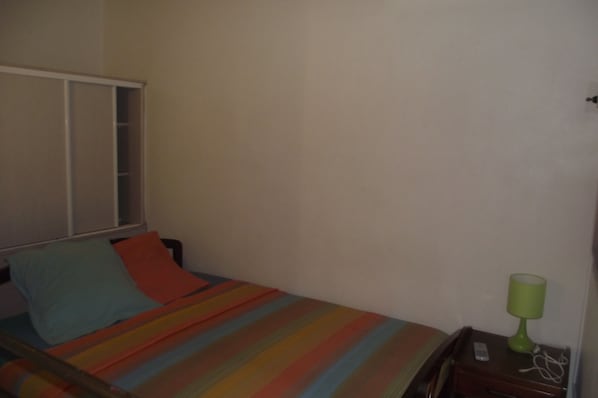 A single bed with a colorful striped bedspread, a green bedside lamp, and a window with closed blinds.