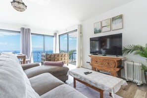 Ein geräumiges Wohnzimmer mit einem großen Flachbildfernseher, einem bequemen Sofa, einer Holzkommode und Meerblick durch große Fenster.