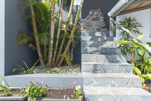Eine Treppe mit Steinstufen führt zu einer Steinmauer, umgeben von verschiedenen Pflanzen und einer kleinen Felsformation.