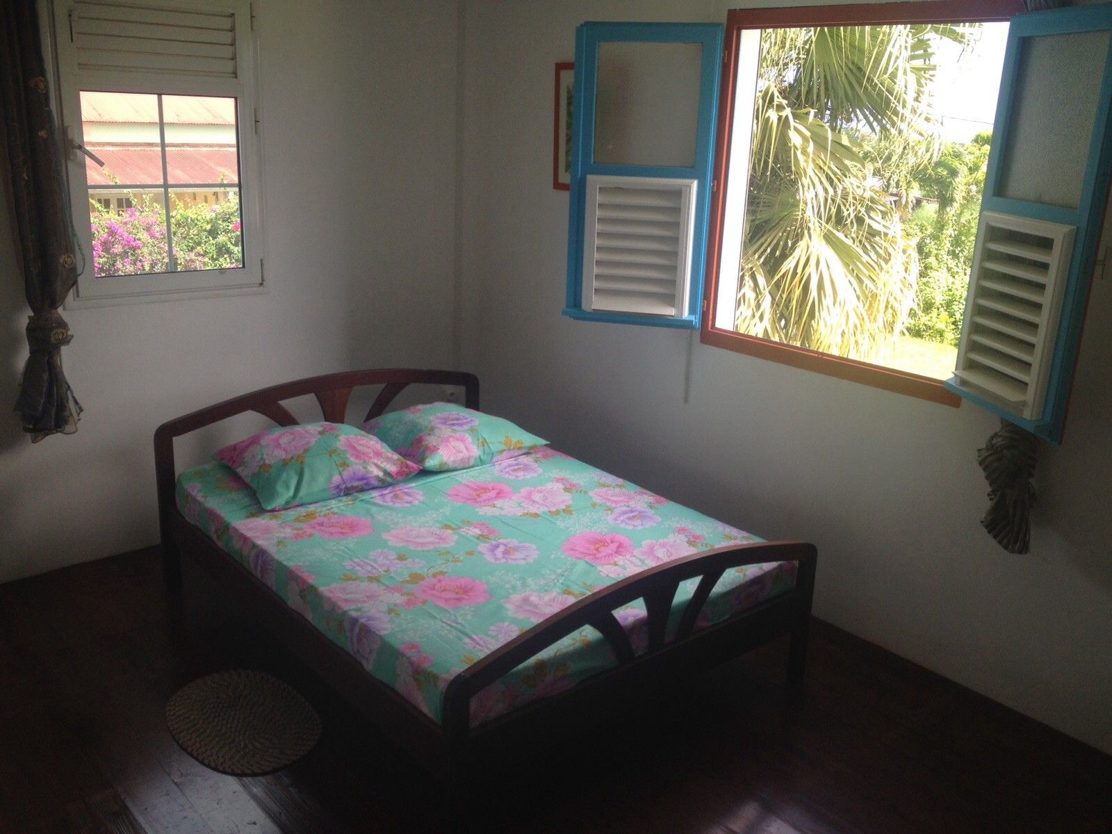 Una habitación pequeña con una cama individual, estructura de madera, colcha estampada con motivos florales, ventana con contraventanas y vista a zonas verdes.