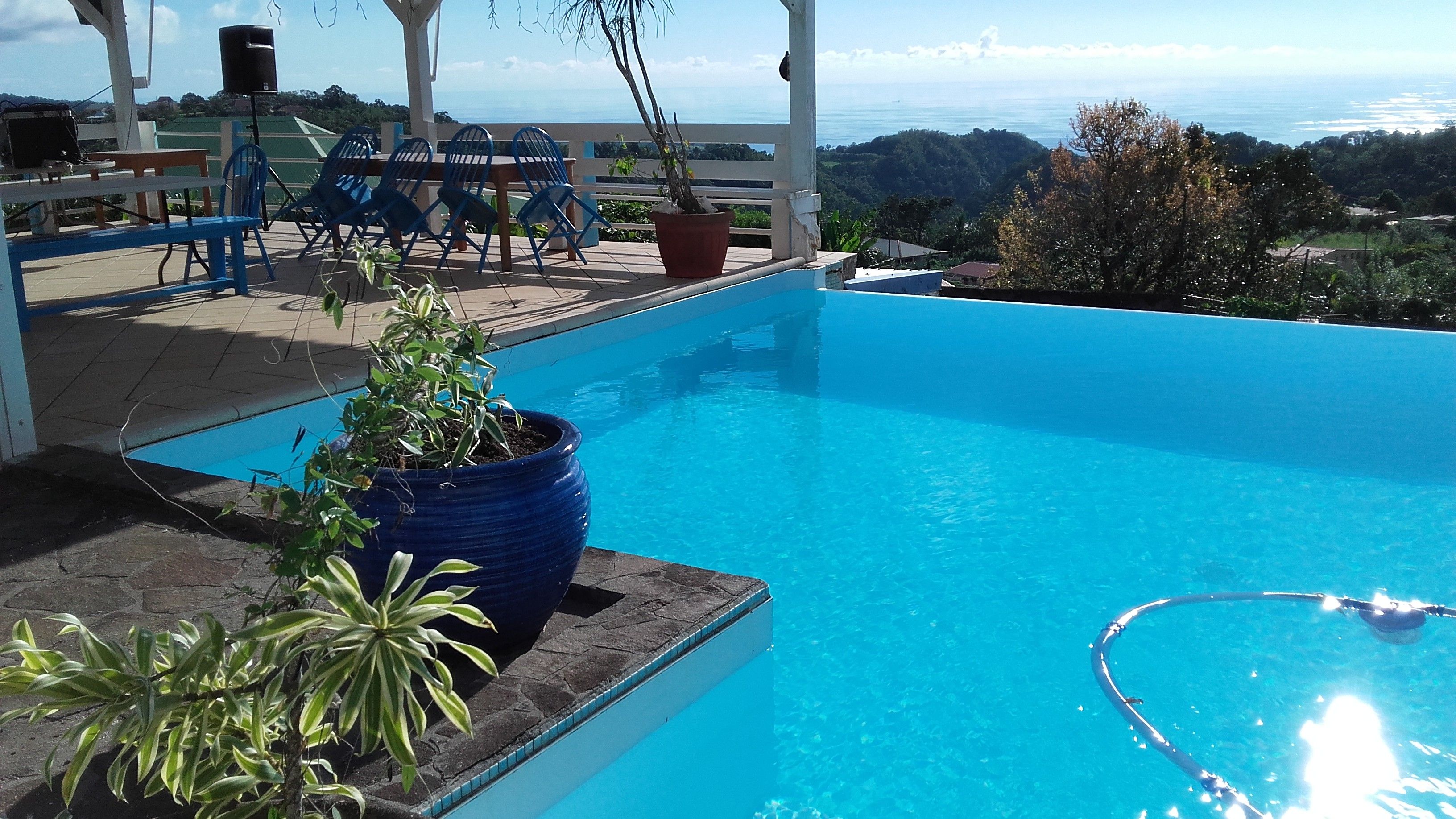 Une piscine d’un bleu éclatant, avec vue sur l’océan et les collines, entourée de chaises et d’une table, toutes de couleur bleue.