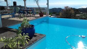 Une piscine d’un bleu éclatant, avec vue sur l’océan et les collines, entourée de chaises et d’une table, toutes de couleur bleue.