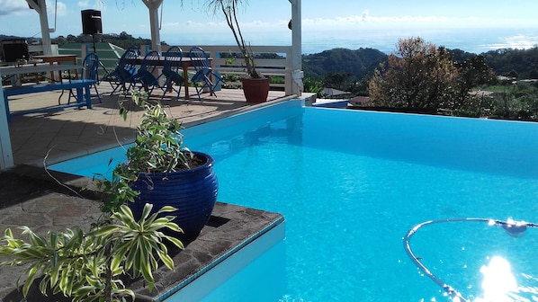 A clear blue pool with a view of the ocean and hills, surrounded by blue chairs and a table.