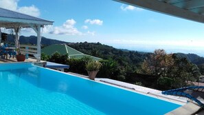 Piscina azul e límpida com vista para uma colina verde e o oceano ao fundo.