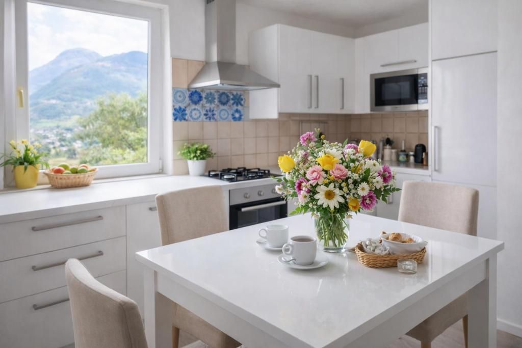 Une cuisine moderne avec des meubles blancs, une cuisinière et un four à micro-ondes. On y trouve une table à manger avec des chaises, un vase de fleurs et, par la fenêtre, une vue sur les montagnes.