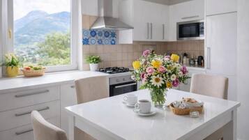Une cuisine moderne avec des meubles blancs, une cuisinière et un four à micro-ondes. On y trouve une table à manger avec des chaises, un vase de fleurs et, par la fenêtre, une vue sur les montagnes.