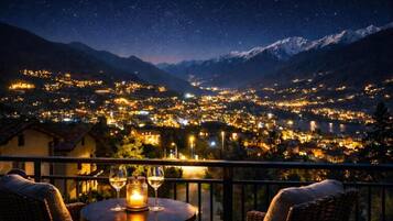Vista nocturna de un pueblo con una montaña de fondo, un balcón con muebles de mimbre y una mesa con dos copas de vino y una vela.
