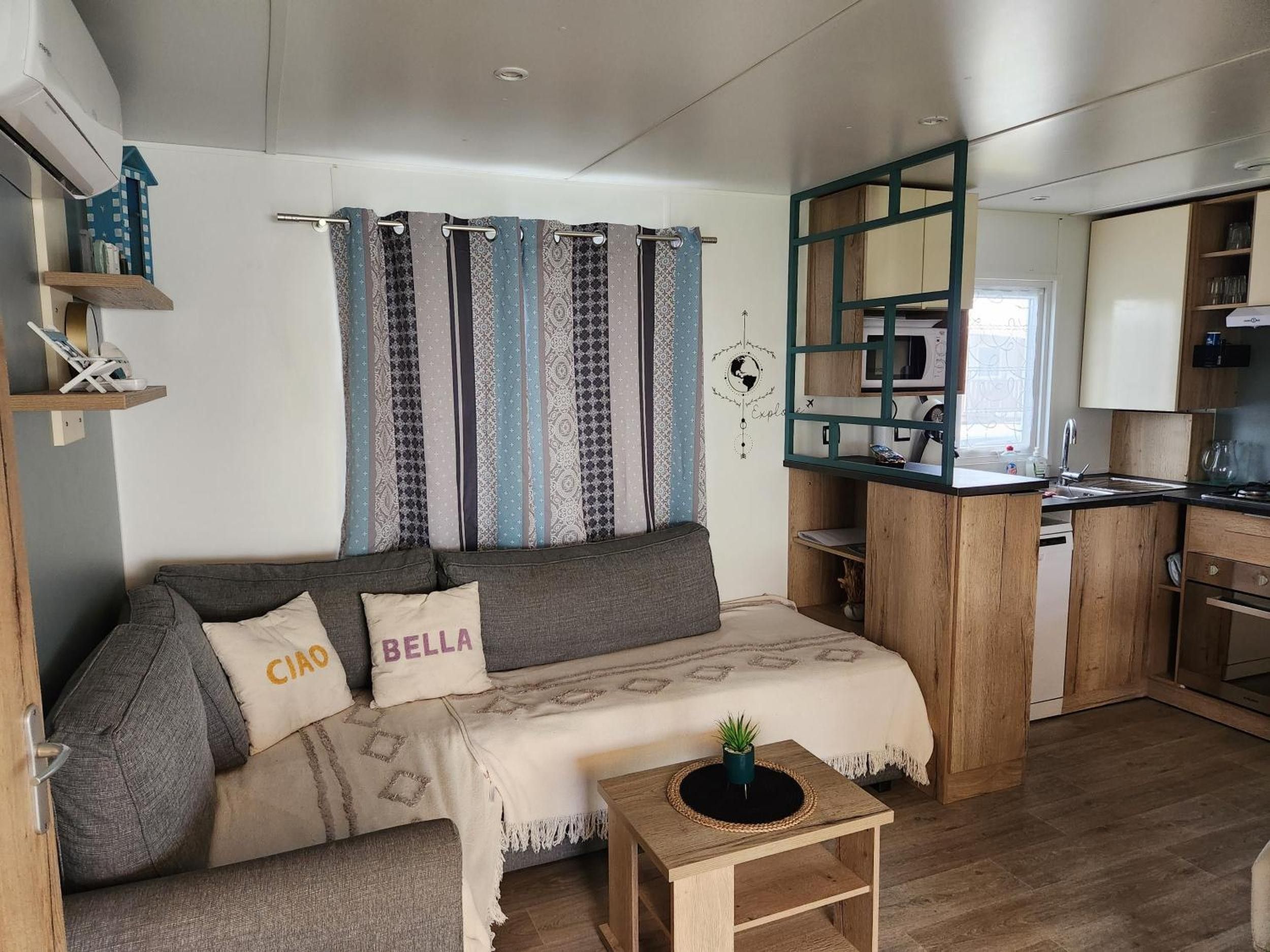 Ein gemütlicher Wohnbereich mit einer Couch, einem kleinen Beistelltisch, einer Küchenzeile mit Mikrowelle und einem Fenster mit Vorhängen.