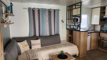Ein gemütlicher Wohnbereich mit einer Couch, einem kleinen Beistelltisch, einer Küchenzeile mit Mikrowelle und einem Fenster mit Vorhängen.