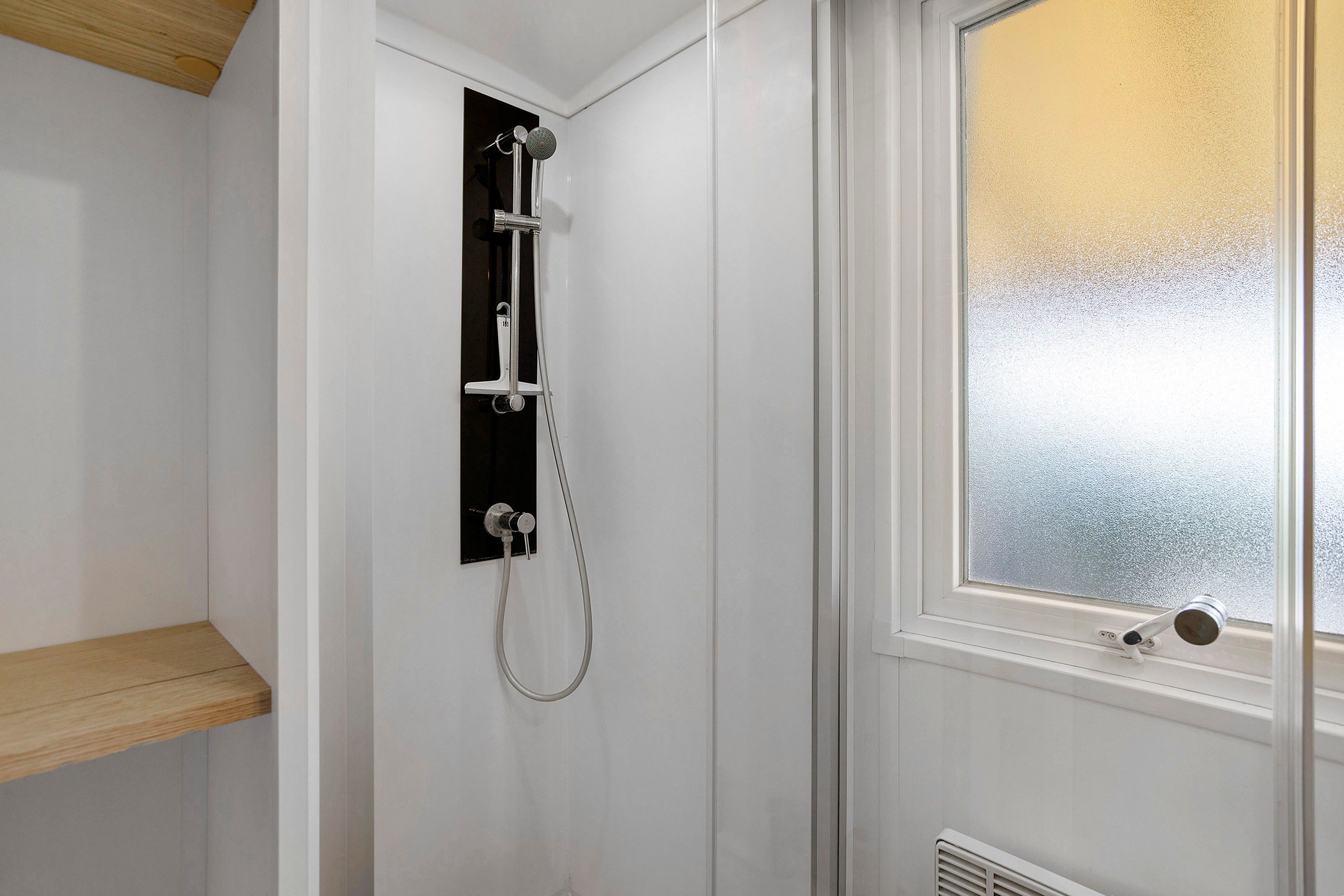 A shower with a glass door, a wooden shelf, and a window with frosted glass.