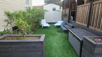 A backyard with artificial grass, two white lounge chairs, wooden planters, and a small shed.