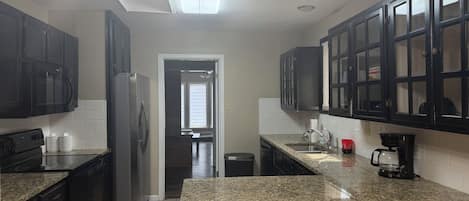 A kitchen with granite countertops, black cabinets, a stainless steel refrigerator, a coffee maker, and a trash can.
