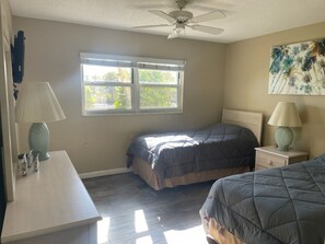 Quarto com duas camas, um ventilador de teto, janela com persianas, mesa de cabeceira com abajur e uma obra de arte na parede.
