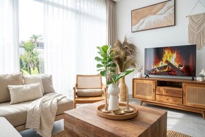 Ein modernes Wohnzimmer mit einer Couch, einem Couchtisch mit Dekoration, einem Fernseher, der ein Feuer zeigt, und einem großen Fenster mit Vorhängen.