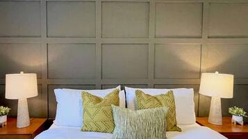 A neatly made bed with white bedding and multiple pillows, flanked by two bedside tables with lamps and small potted plants.