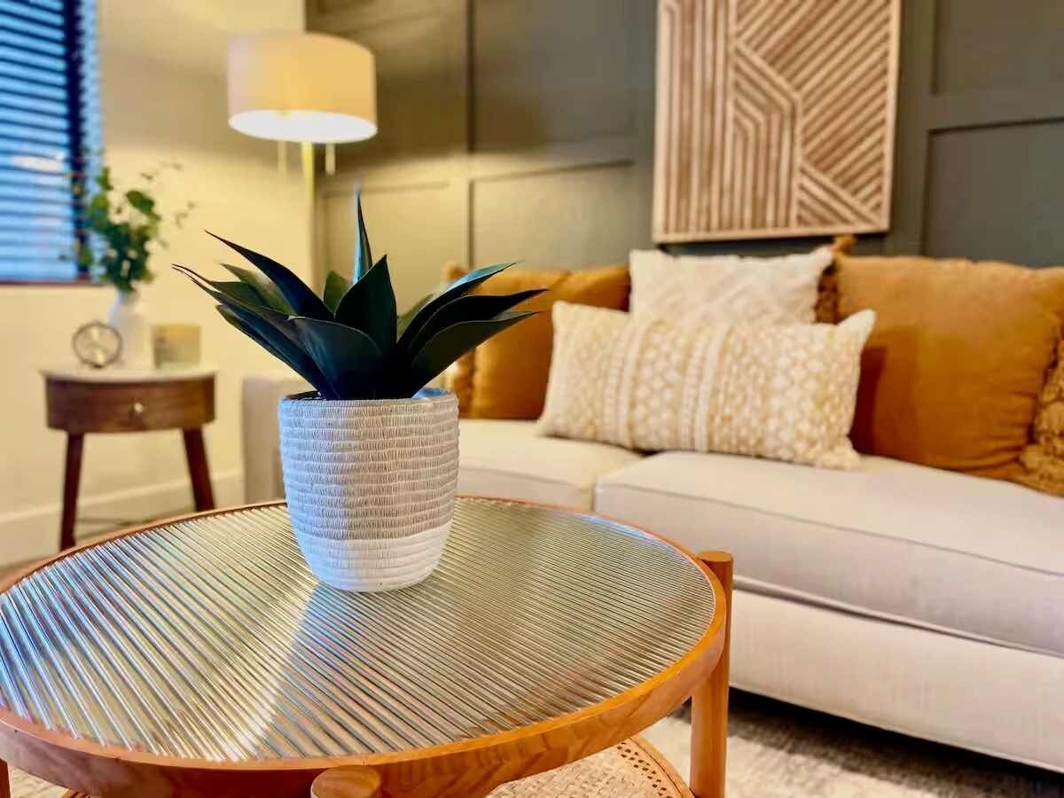 A round table with a woven texture, a potted plant, and a white sofa with patterned cushions in a living room.