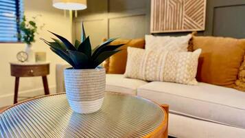 A round table with a woven texture, a potted plant, and a white sofa with patterned cushions in a living room.
