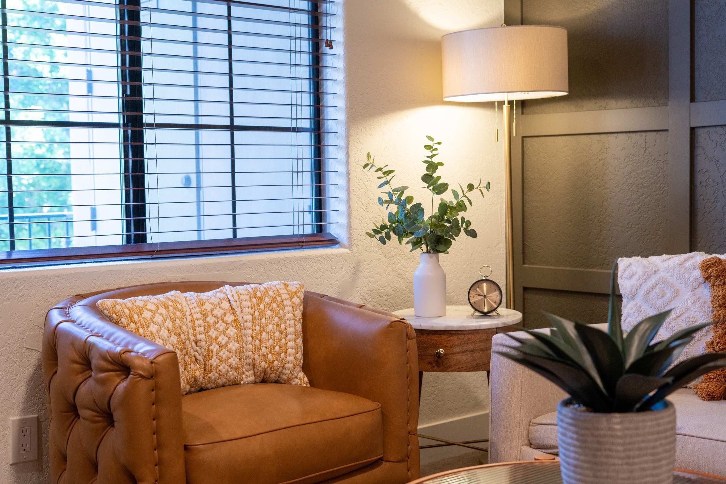 A cozy living room with a leather armchair, a side table with a lamp, a clock, and a potted plant.