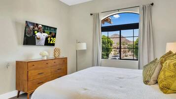 A bedroom with a bed, a dresser, a television displaying a tennis player, and a window with a view of the outdoors.