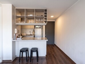 A compact kitchen with a countertop, two stools, and a built-in shelving unit above the sink. - Edificio Zañartu by Domus Travels (Ñuñoa)