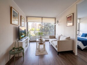 Ein modernes Wohnzimmer mit einer Couch, einem Fernseher, einem kleinen Tisch mit Pflanzen und Blick auf die Stadt durch große Fenster.