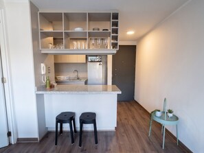Eine kompakte Küche mit integriertem Kühlschrank, einer Spüle und einer Arbeitsplatte. Zwei Hocker und ein kleiner Tisch mit Pflanzen sind ebenfalls vorhanden.