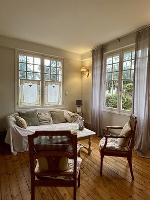 Una habitación con piso de madera, un sofá, dos sillas, una mesita y una lámpara. Hay ventanas con cortinas y vistas a follaje exterior.