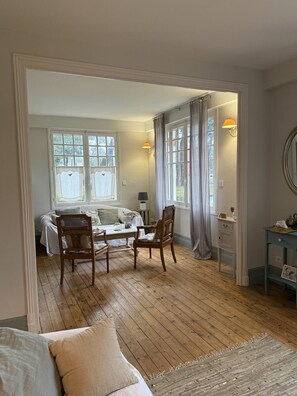 A room with a bed, two chairs, a table, and a small table with a lamp. There are windows with curtains and a view outside. - "The Squirrels of the Park" 400 meters from the beach (Royan)