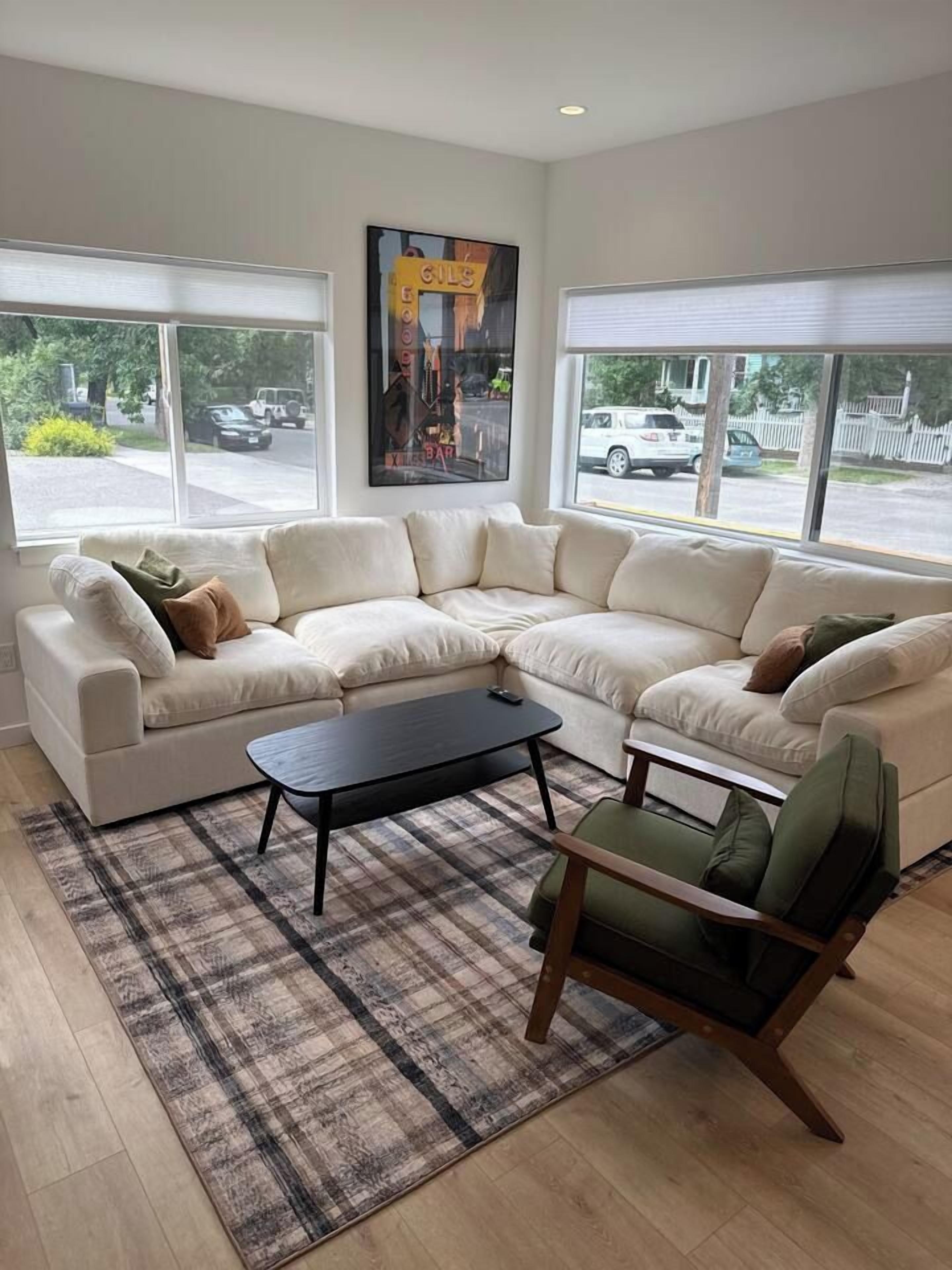 A modern living room with a sectional sofa, a coffee table, and a chair. There are large windows providing a view of the street.