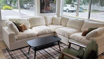 A modern living room with a sectional sofa, a coffee table, and a chair. There are large windows providing a view of the street.