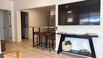 A modern living room with a wooden dining table and bar stools, a black console table with decorative items, and a large flat-screen TV mounted on the wall.