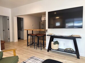 A modern living room with a wooden dining table and bar stools, a black console table with decorative items, and a large flat-screen TV mounted on the wall. - Luxury Livingston Retreat + Hot Tub (Livingston)