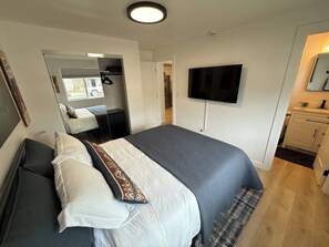 A hotel room with a bed, a flat-screen TV mounted on the wall, a mirror, and a small kitchenette area.