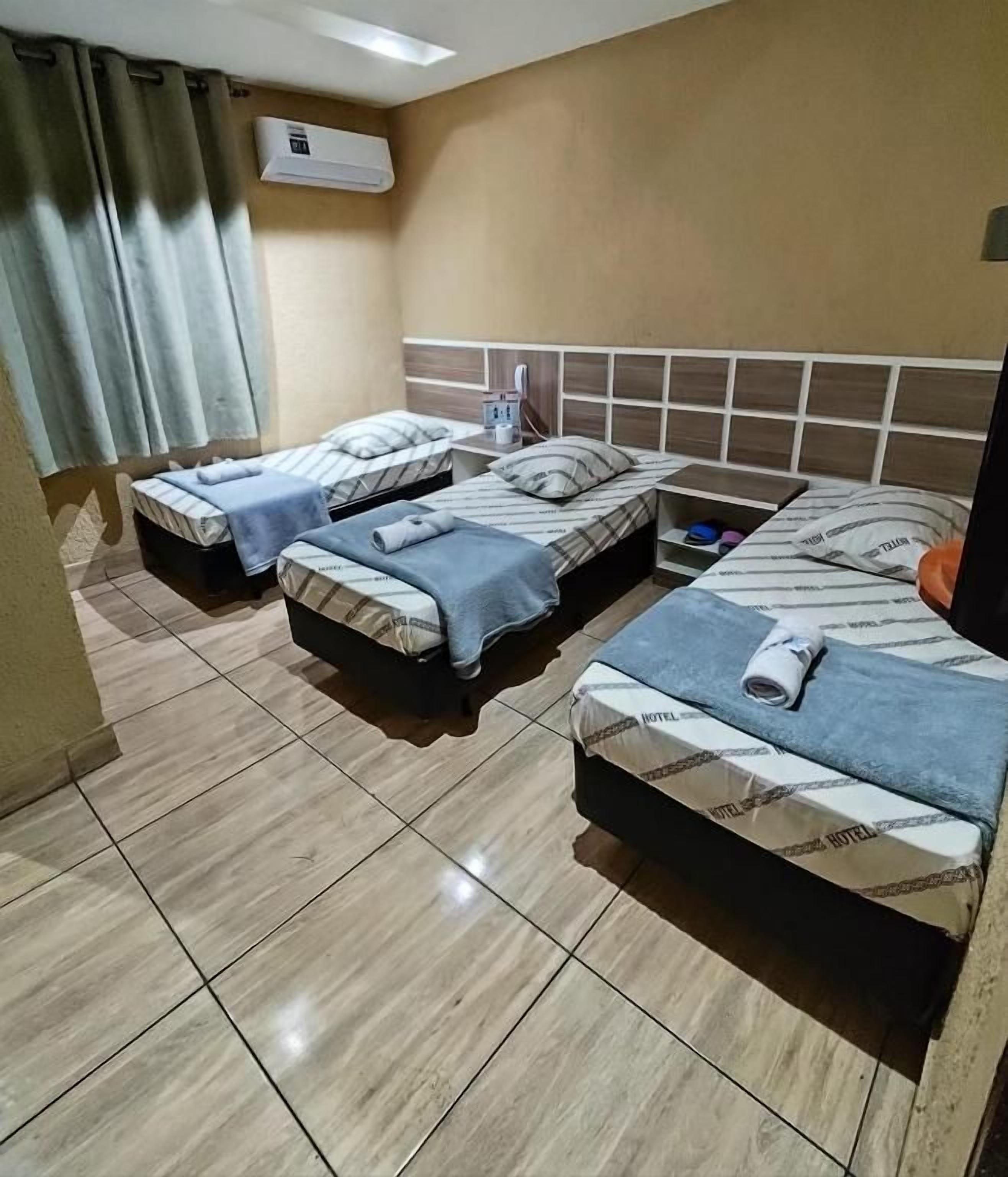 Habitación de hotel con tres camas, una ventana con cortinas, un equipo de aire acondicionado y piso de baldosas.