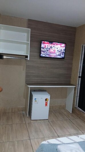 Habitació senzilla amb televisió penjada a la paret, nevera petita i armari blanc.