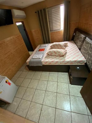 Habitación pequeña y acogedora con cama, mesita de noche, televisor montado en la pared y ventana con cortinas.