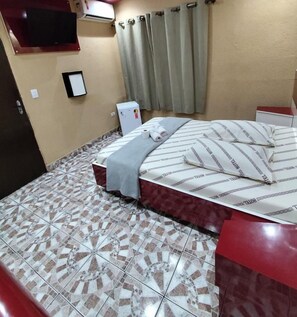Habitació d'hotel amb un llit, una televisió penjada a la paret, un frigorífic petit i una finestra amb cortines.