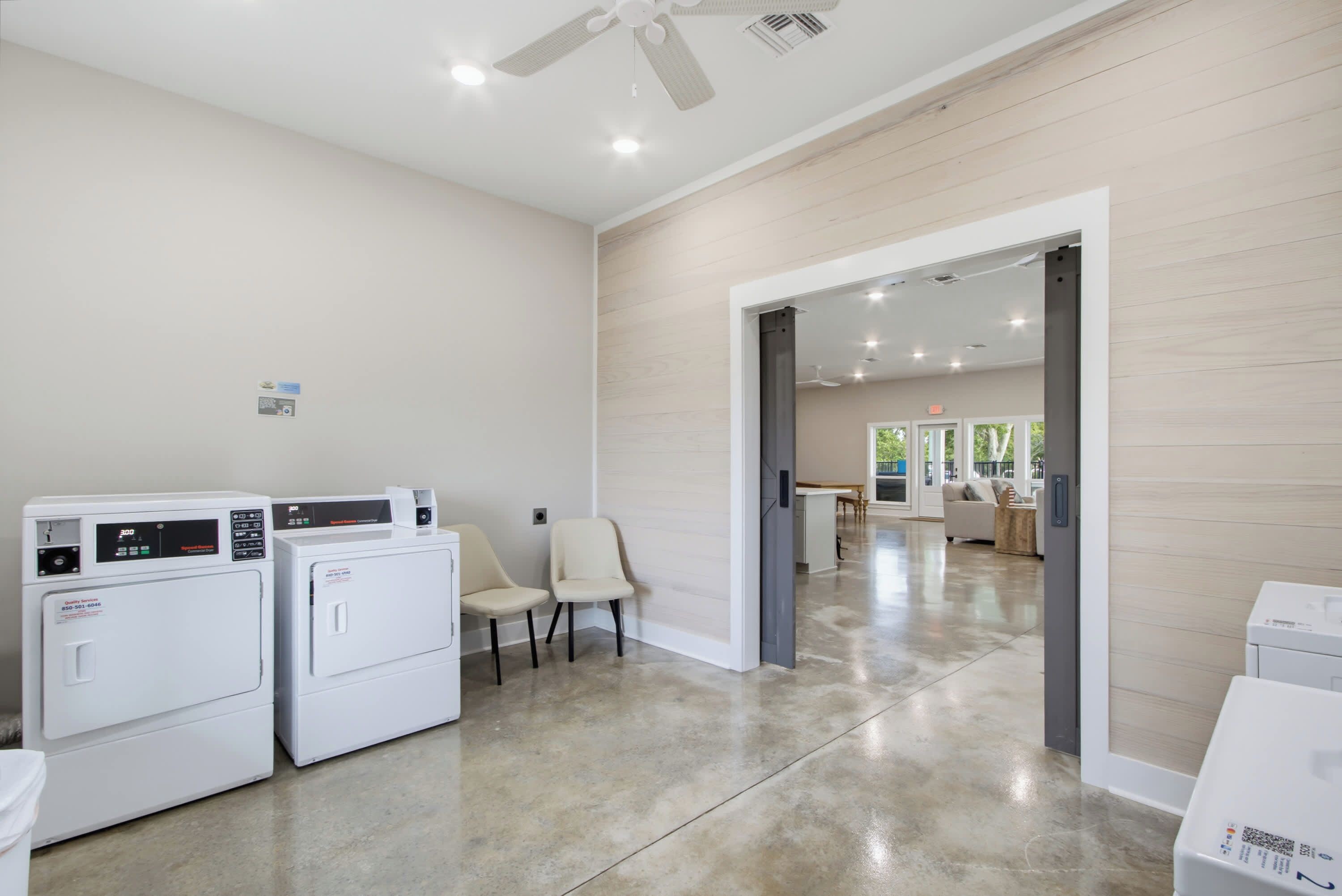 A laundry area with two washing machines, a chair, and a door leading to a living room.