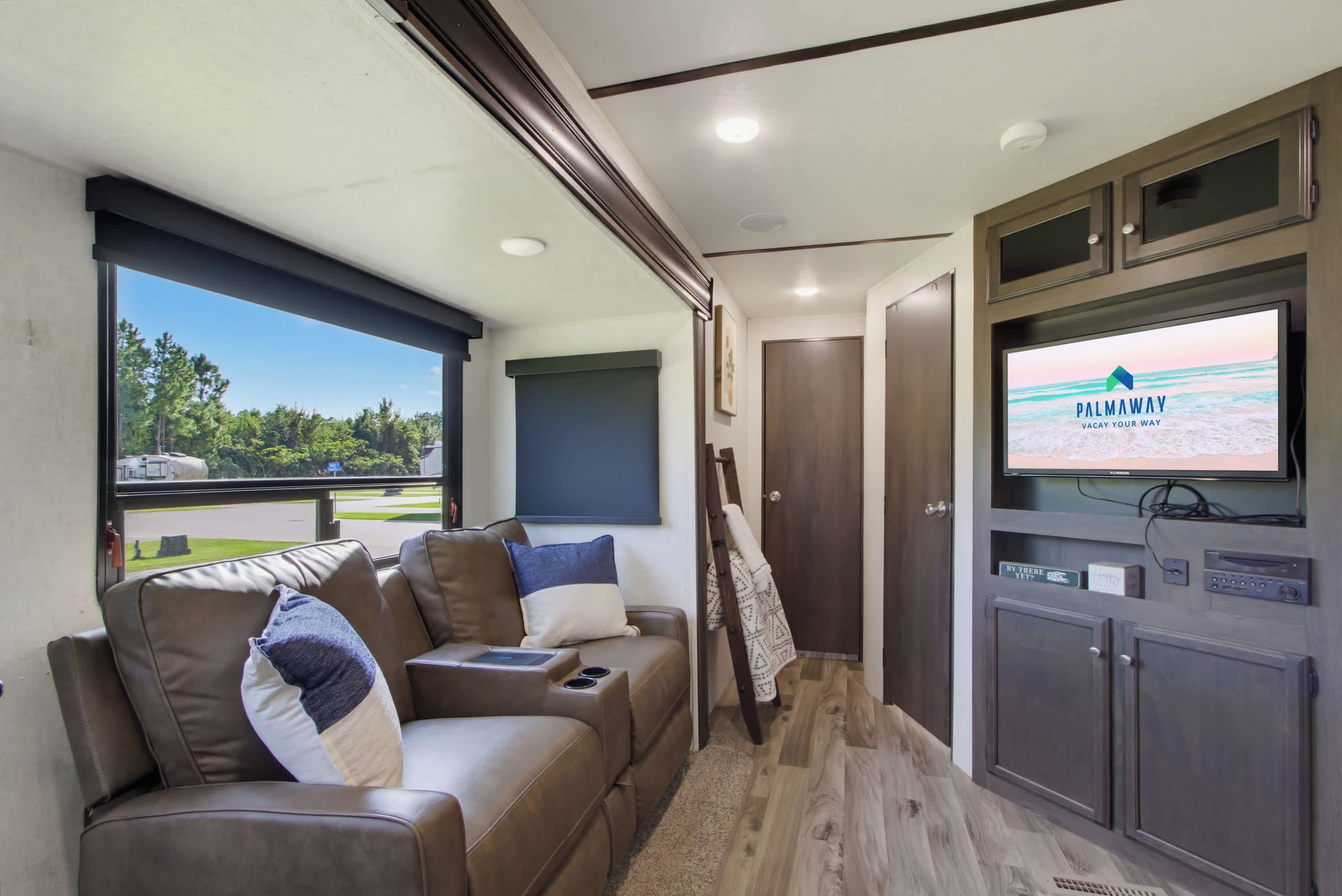 A modern RV interior with a sofa, television, and a view of the outdoors.