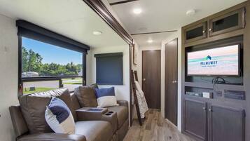 A modern RV interior with a sofa, television, and a view of the outdoors.