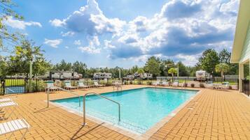 A swimming pool with a clear blue sky and clouds above, surrounded by lounge chairs and RVs.