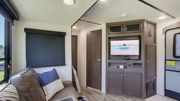 A compact RV interior with a seating area, a mounted TV, and a window providing a view outside.
