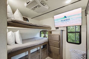 Una cama litera con un televisor mostrando 'PALMAWAY VACAY YOUR WAY' y una ventana con vista a follaje.