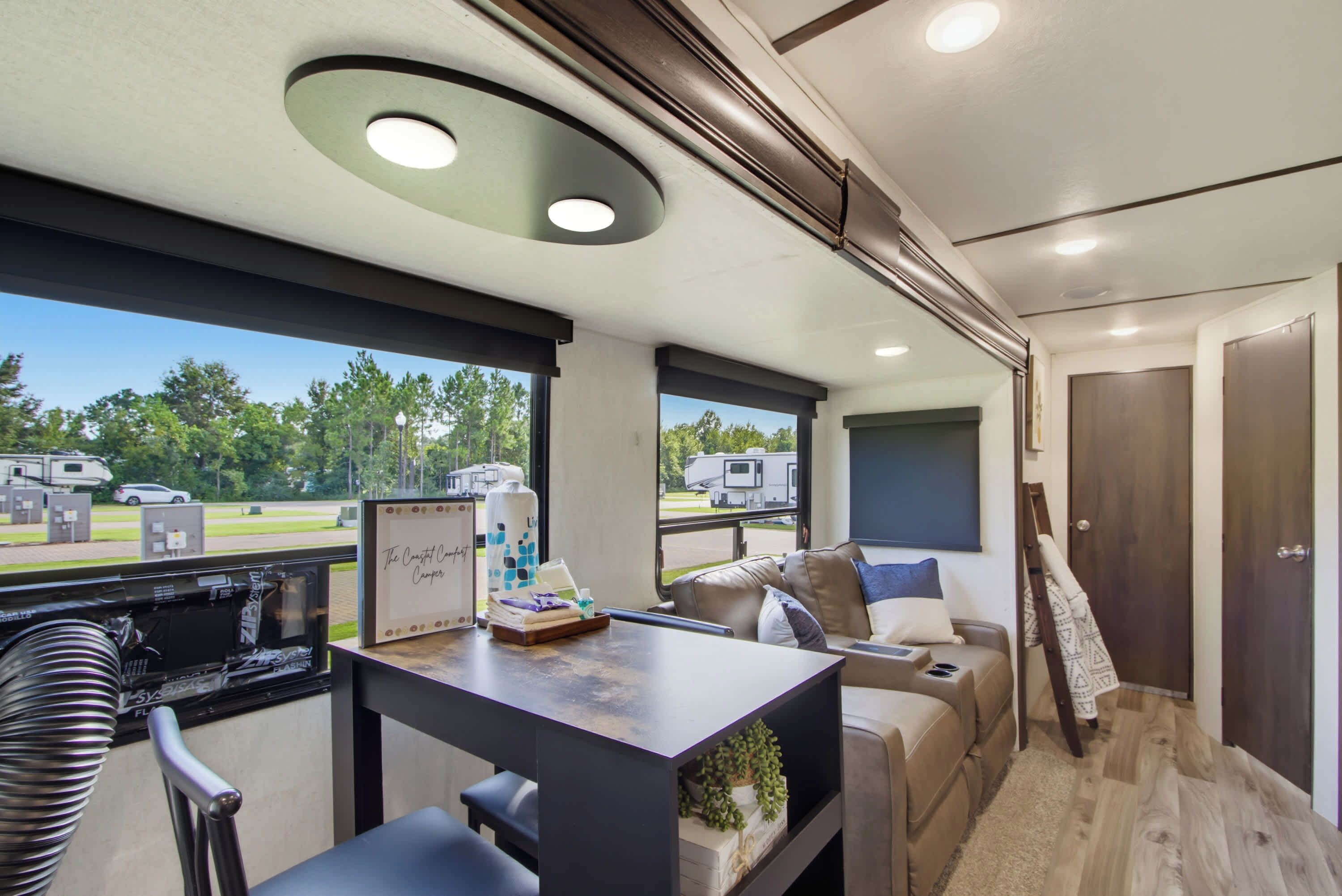 A modern RV interior with a dining area, seating, and a view of the outdoors.