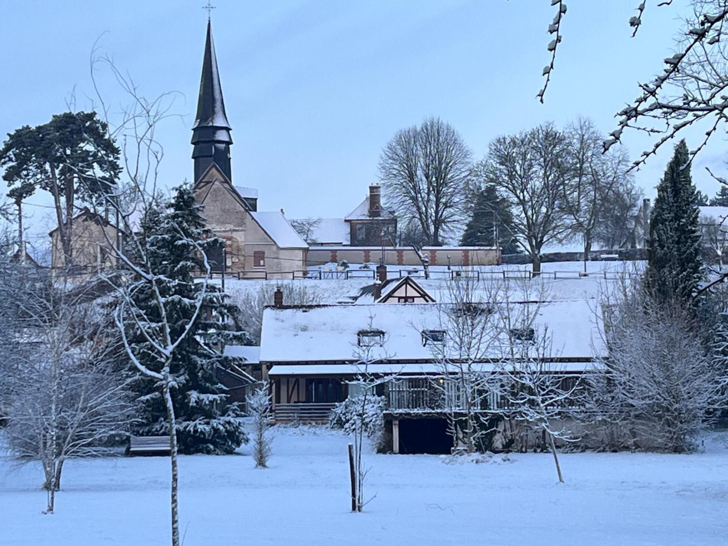 Una chiesa innevata con un campanile, circondata da alberi spogli e un edificio in legno.