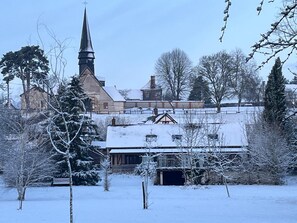 Eine schneebedeckte Kirche mit einem Kirchturm, umgeben von kahlen Bäumen und einem Holzhaus.